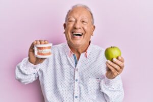 Man smiling while holding his dentures and an apple